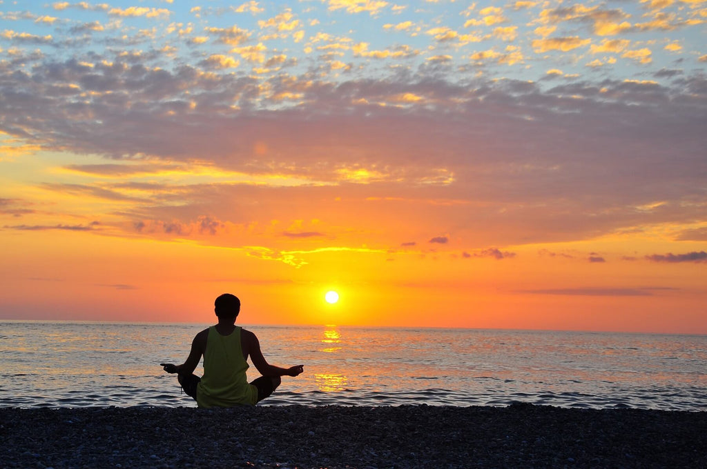 Man in lotus position on beach facing golden sunset